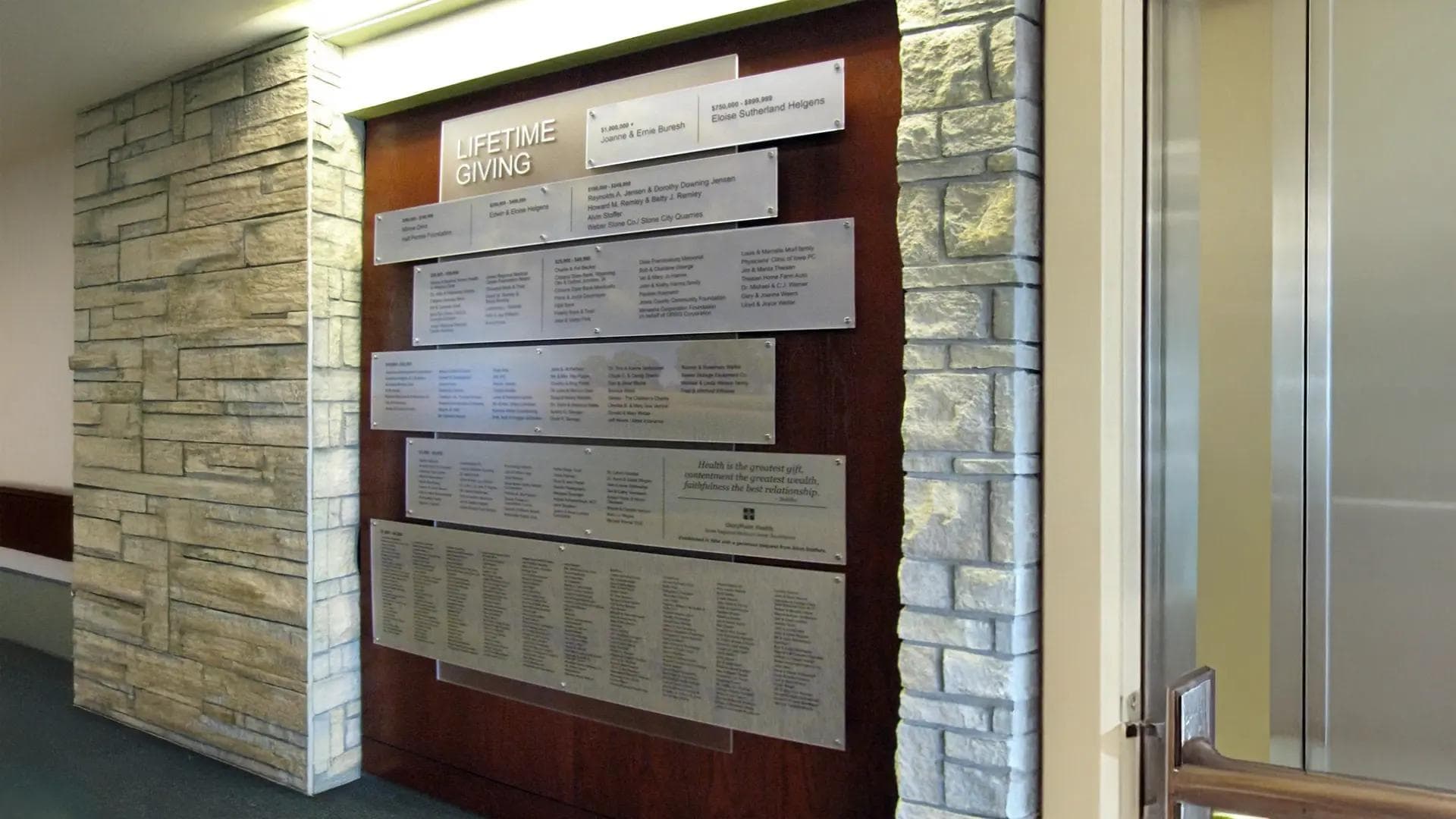 Lifetime Giving donor wall with layered metal panels mounted on a wood backdrop, framed by stone accents in a healthcare corridor.