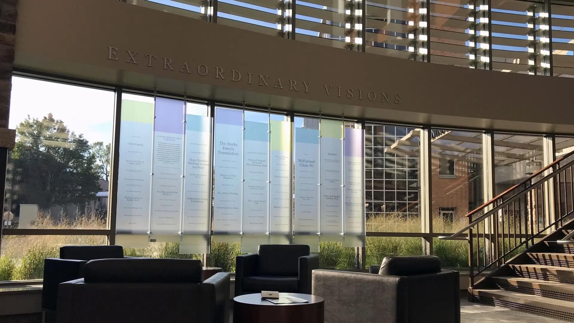 Glass donor recognition wall with suspended panels and “Extraordinary Visions” header in a light-filled hospital lobby.