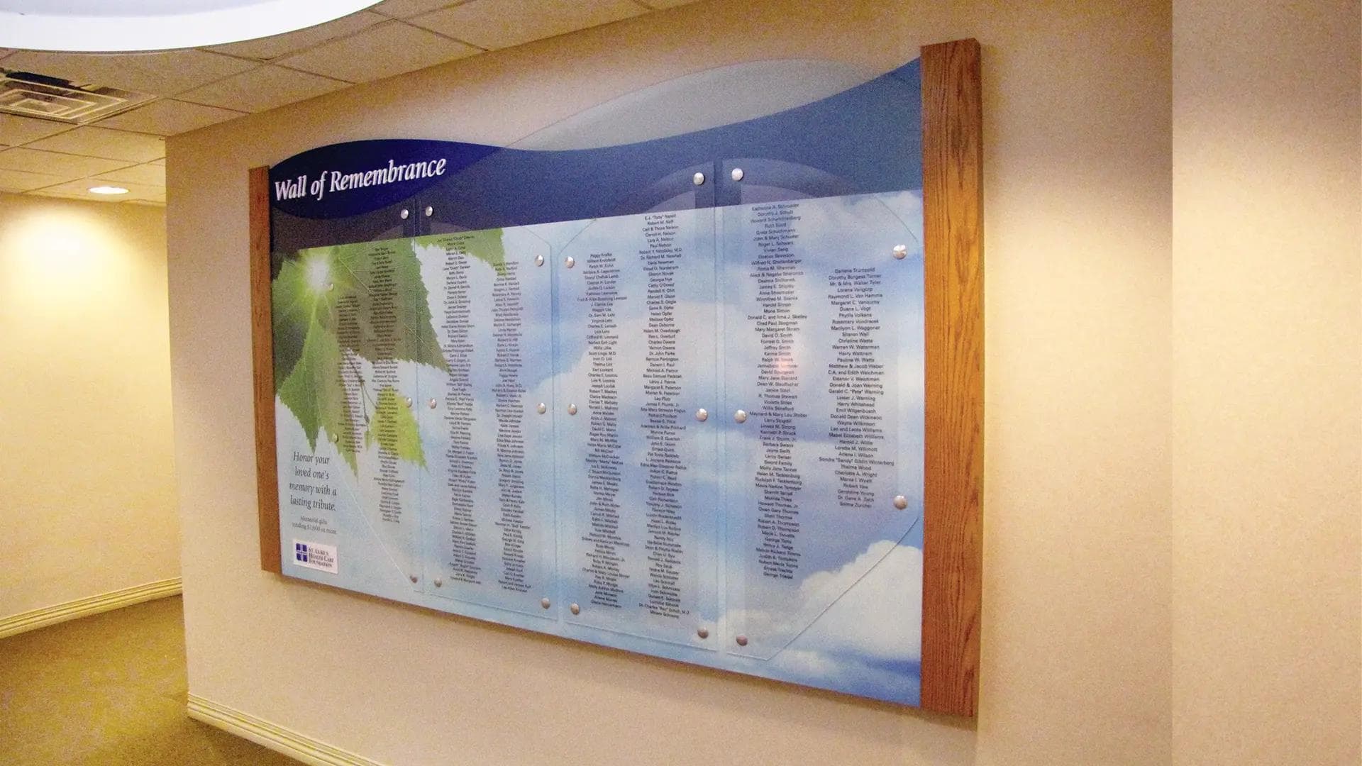 Wall of Remembrance donor wall with printed names on acrylic panels, soft sky imagery, and wood side accents in a quiet corridor.